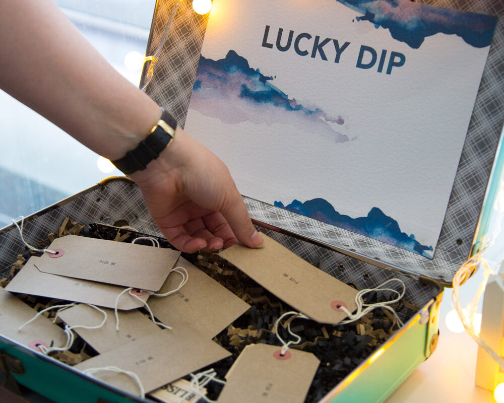 A small vintage suitcase lies open on a ledge. Inside are lots of brown paper luggage tags, and a printed sign that reads Lucky Dip.