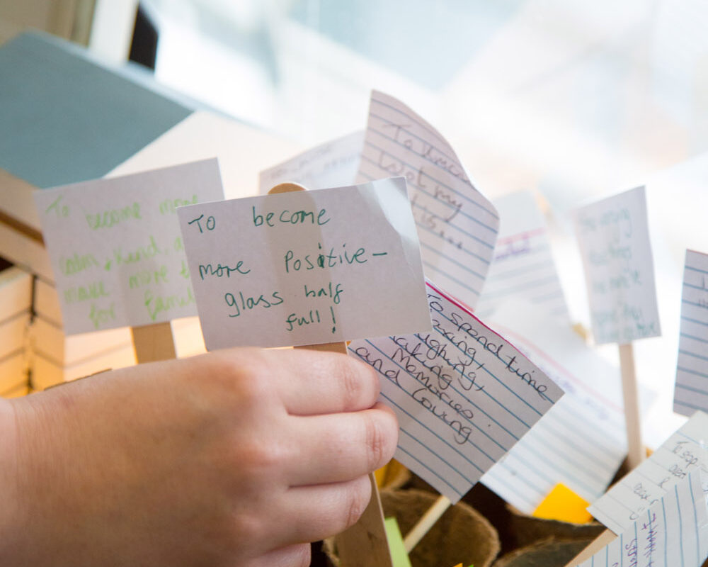 A collection of notes attached to lollipop sticks are standing inside paper cups. A hand adds another note to the collection.