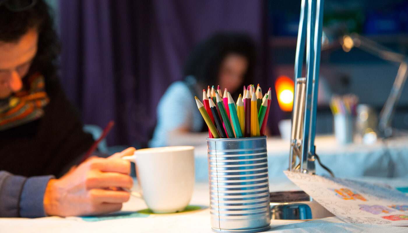 A pot of coloured pencils sits on a table. A woman sits at the table with a mug in her hand.