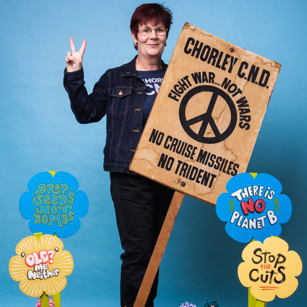 A portrait of a person who holds a placard reading Chorley CND, fight war, not wars. They are holding up two fingers in the peace sign and have placards at their feet that read Old? Me Neither and Stop the Cuts.