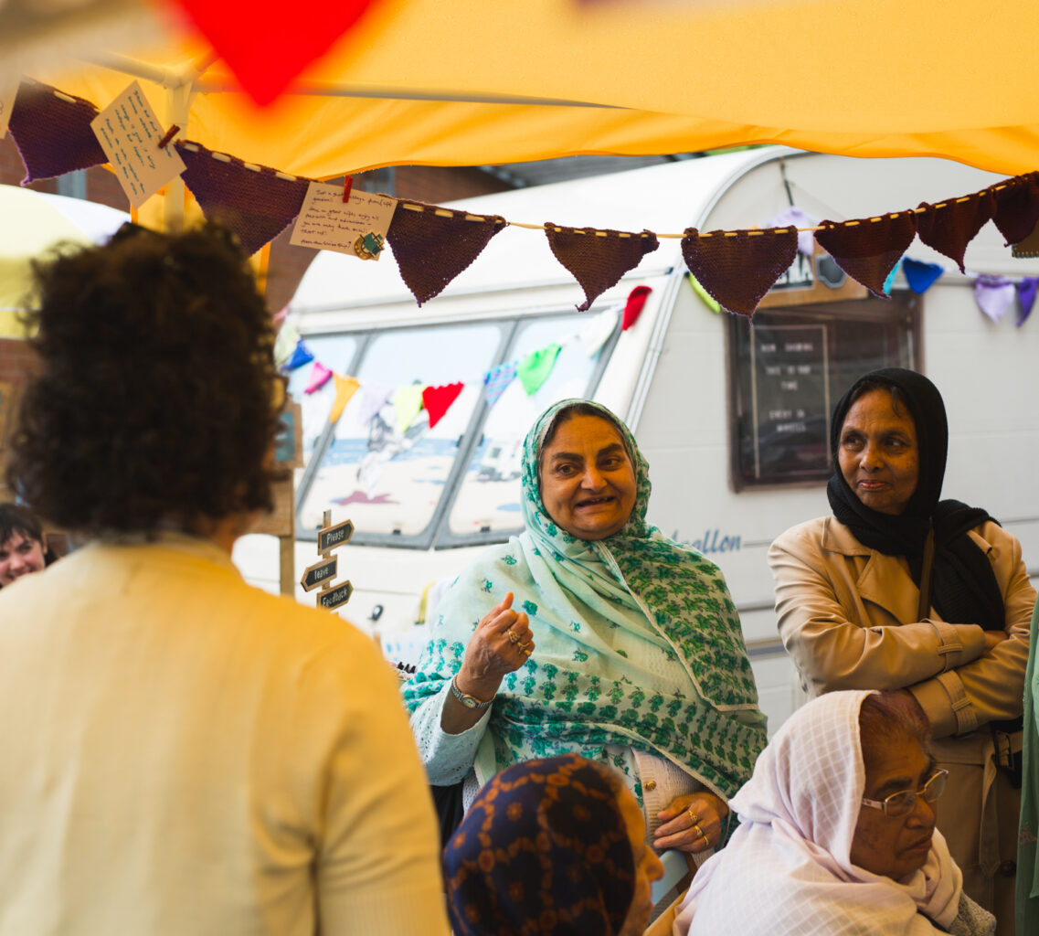 Two women wearing headscarves stand under a marquee. A group is gathered in front of them, and one of the standing women talks to them. In the background there is a caravan.