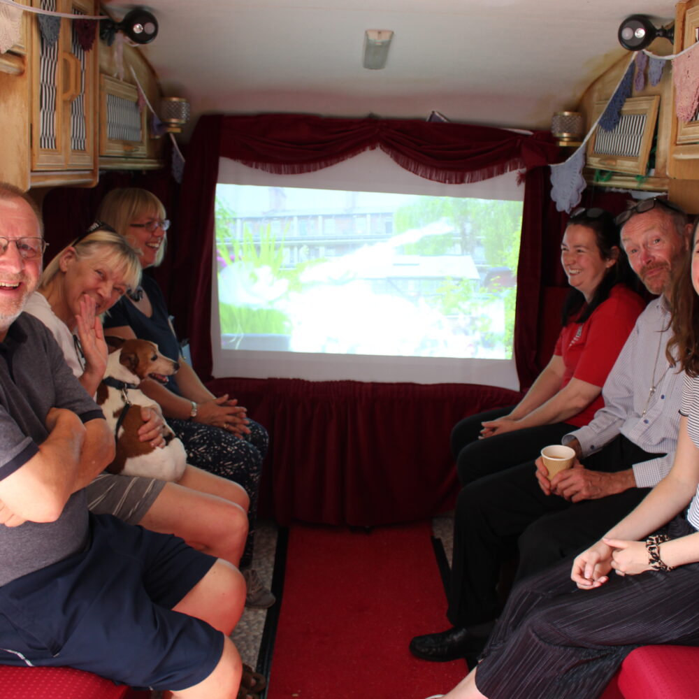 Six adults sit smiling inside a caravan that has red decor, like a cinema. They sit facing each other, in two rows of three. Behind them, a screen is lit up on the back wall.