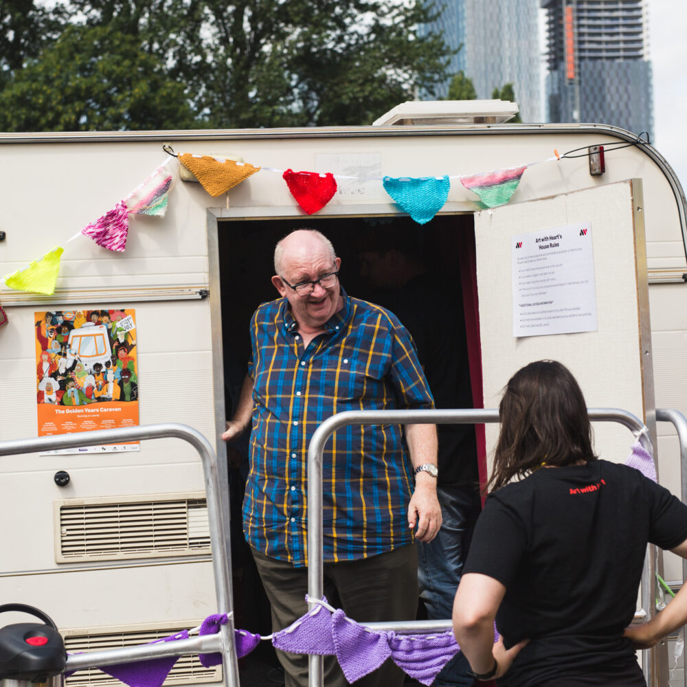 An older person stands in the doorway of a caravan that has been decorated with colourful bunting. They are smiling at someone standing outside the caravan.