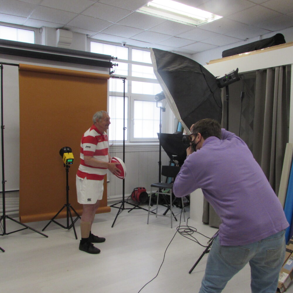 A person with grey hair poses in a photographers' studio. They are wearing a red and white striped sports kit and holding a rugby ball, while a photographer takes their picture.