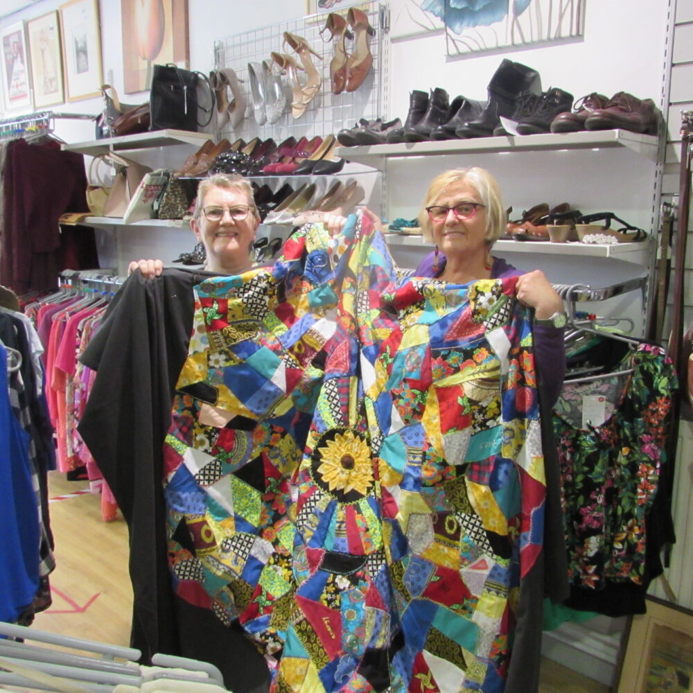 Two people stand next to each other in a charity shop. Together, they are smiling and holding up a large multicoloured patchwork fabric.
