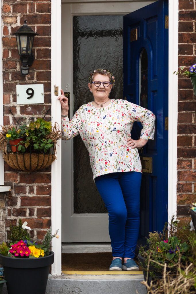 A person poses for a portrait, standing in the doorway of a house with a blue door. They are leaning on the doorframe and smiling widely.