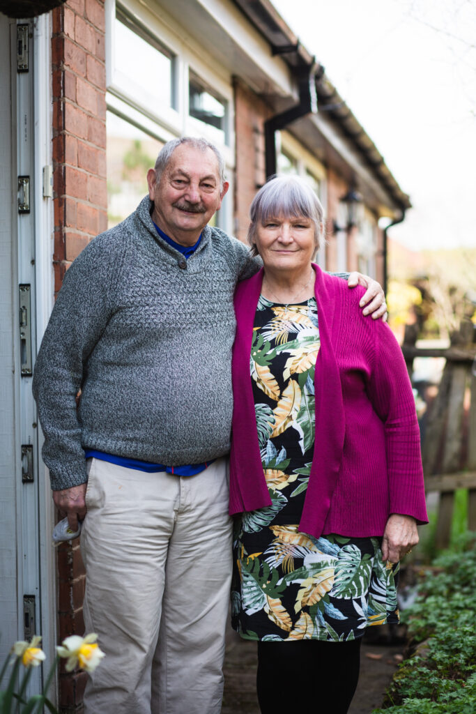 A man and woman pose for a portrait with their arms around each other. They are wearing casual clothes and standing just outside the doorway of a residential property.