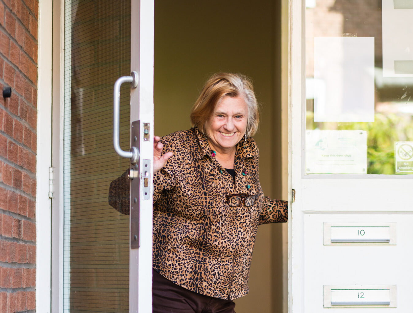 A person wearing a leopard print shirt stand in an open doorway, smiling as though in greeting. To the right of the doorframe, four letterboxes are built into the wall.