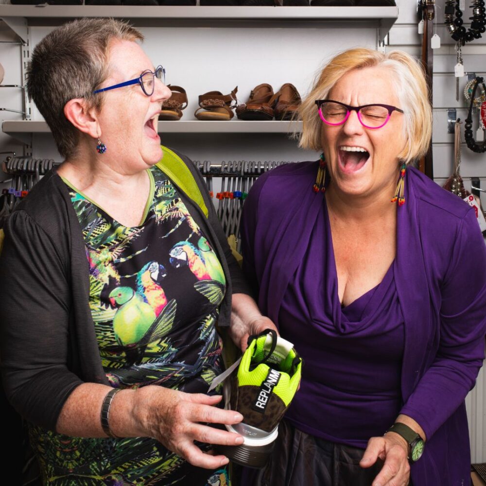 Two people wearing brightly coloured dresses and glasses laugh hysterically together. One of them holds a lime green shoe and behind them is a rack of clothes and shoes.