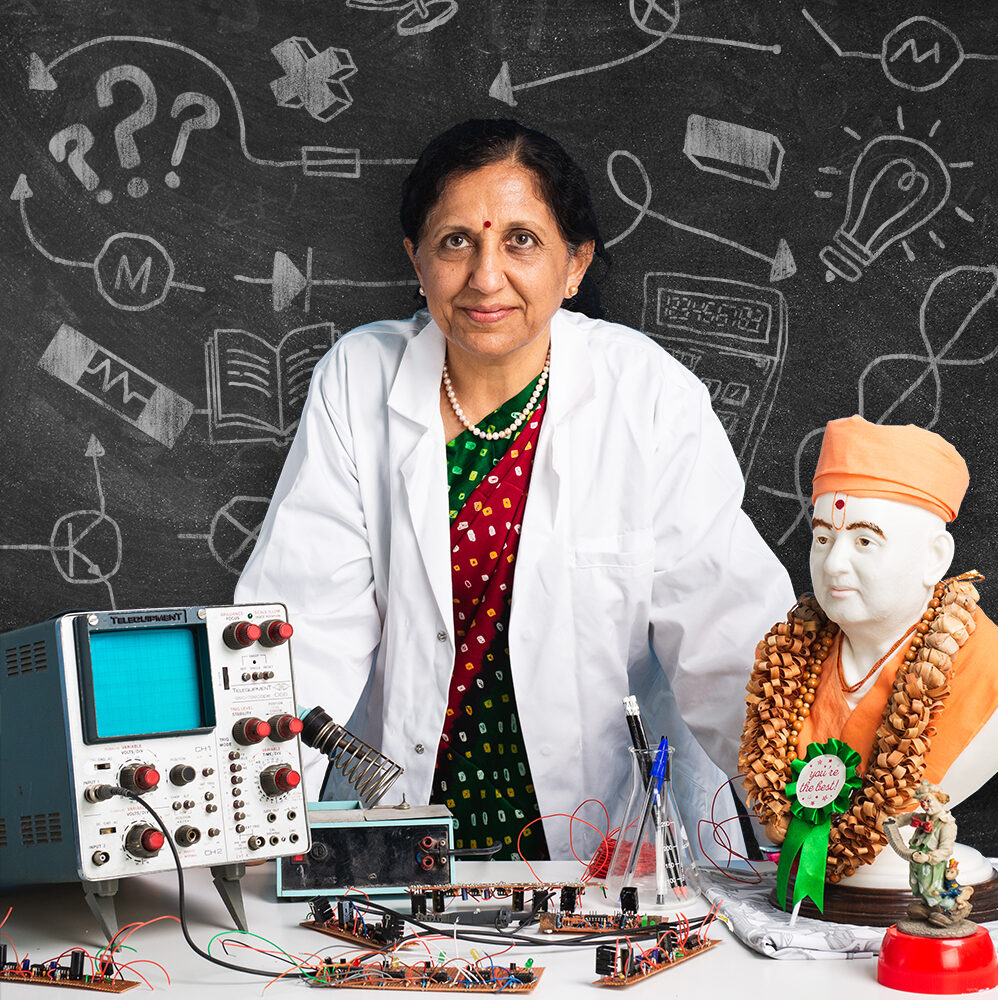 Jagruti is a British asian woman. She is wearing a green and red sari underneath a white lab coat. She is standing behind a desk that is covered with engineering equipment.
