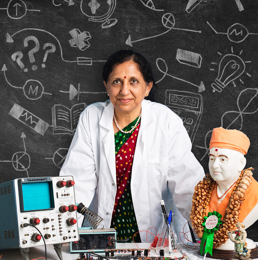 Jagruti is a British asian woman. She is wearing a green and red sari underneath a white lab coat. She is standing behind a desk that is covered with engineering equipment.