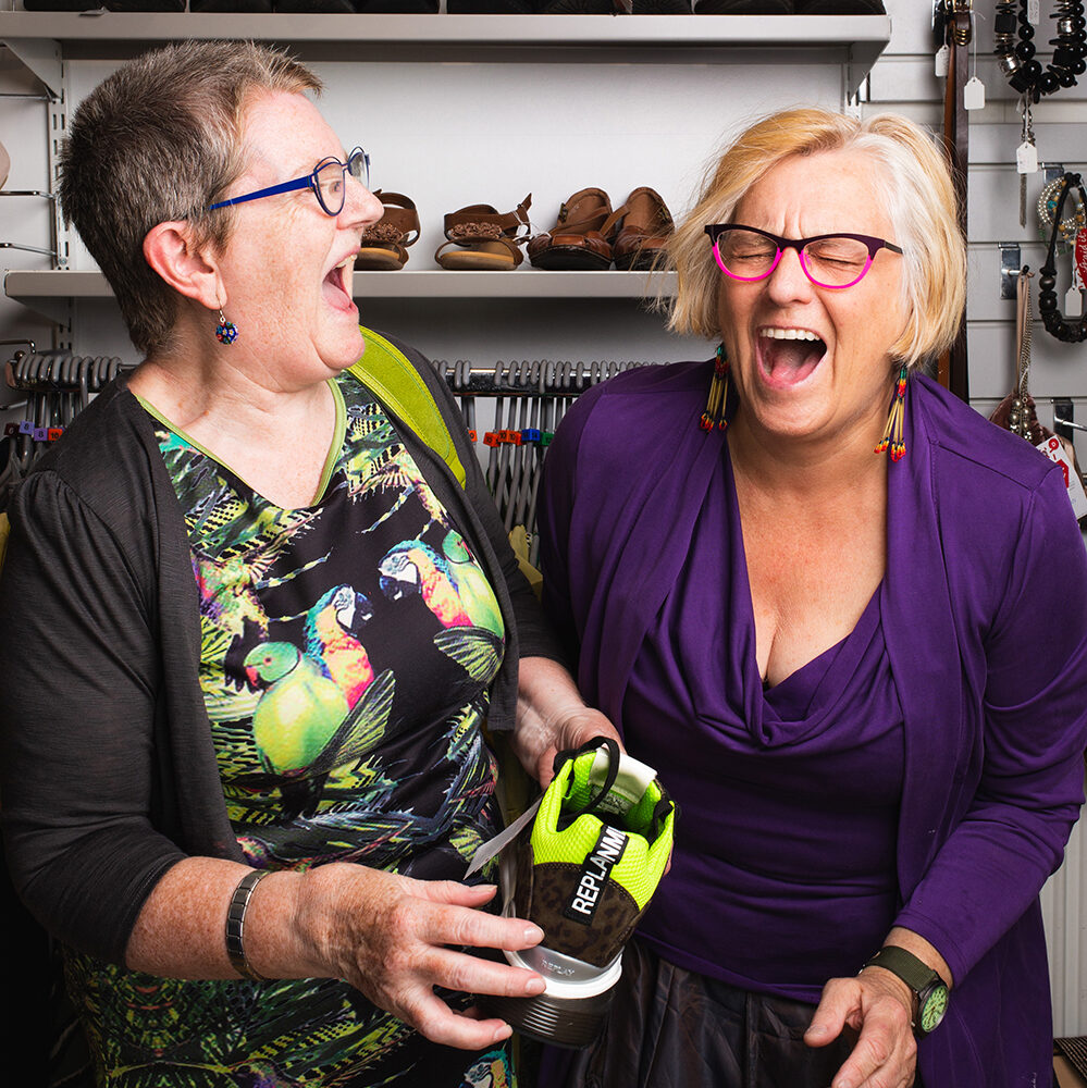 Two women, one with short blonde hair, one with short brown hair are wearing brightly coloured dresses and glasses laugh hysterically together. One of them holds a lime green shoe and behind them is a rack of clothes and shoes.
