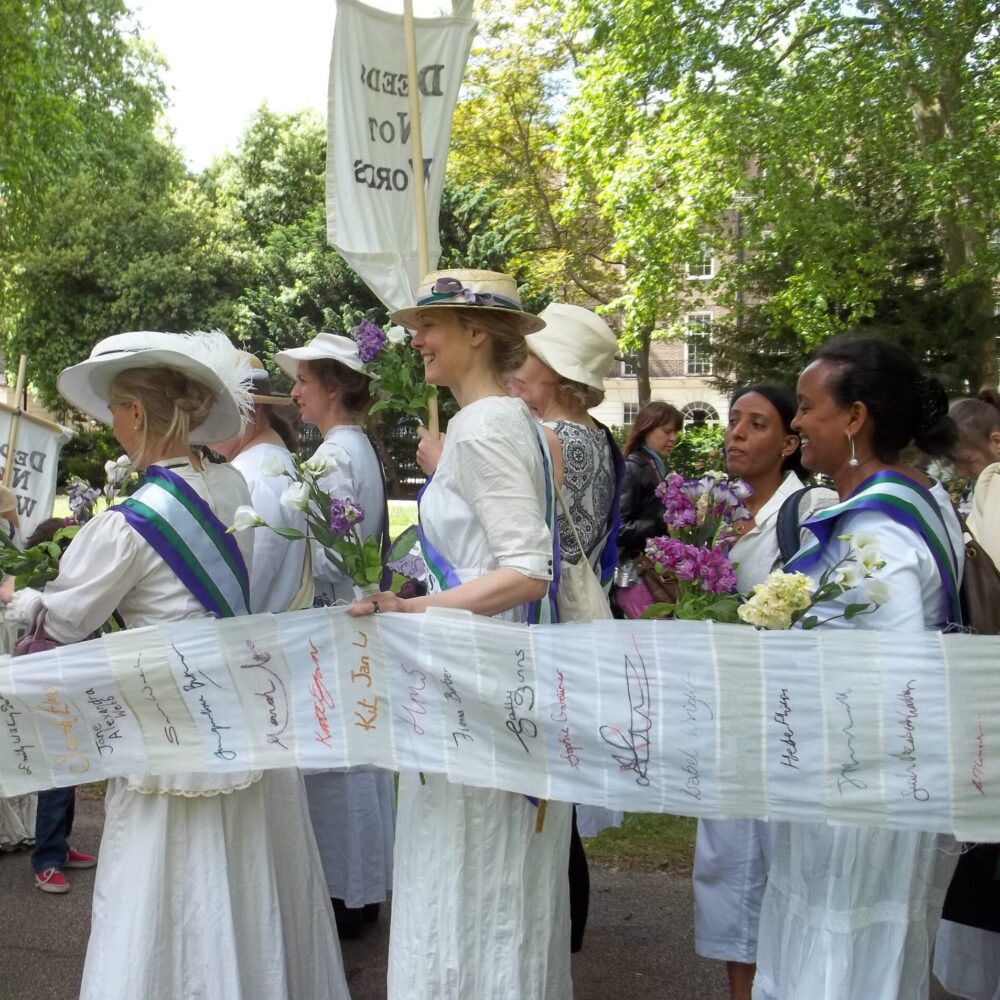 A group of people dressed as suffragettes line up under green trees.