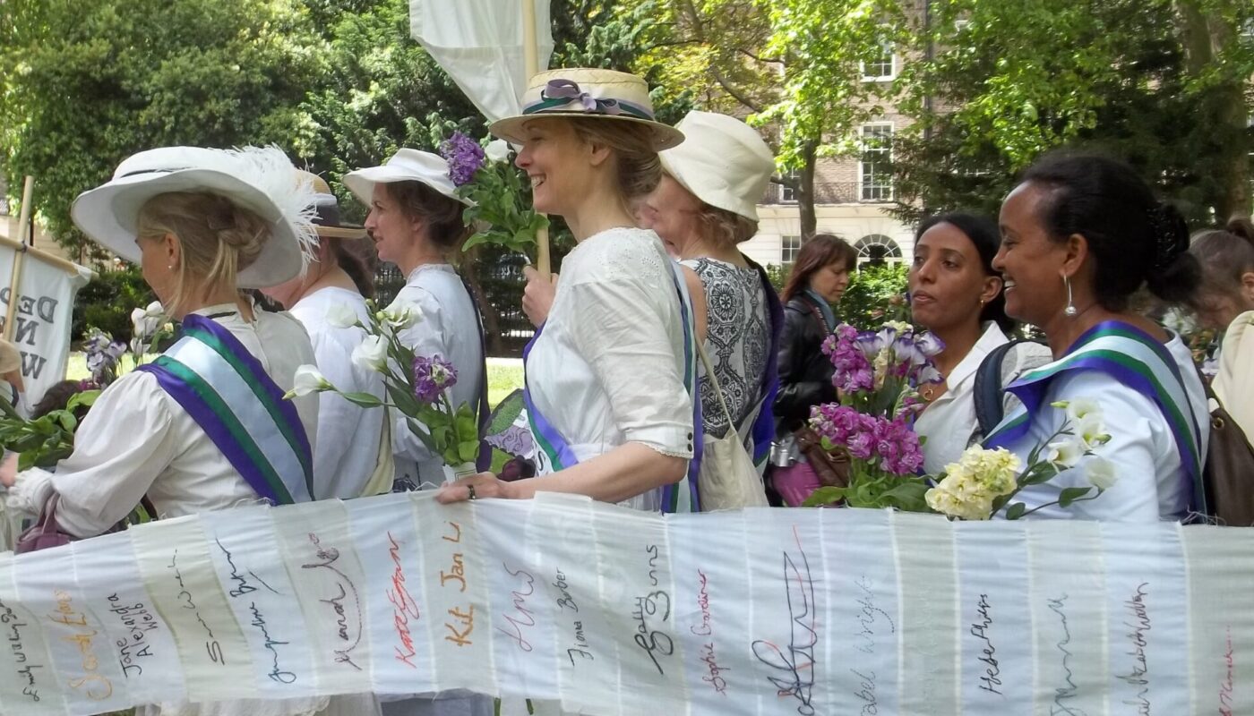 A group of people dressed as suffragettes line up under green trees.