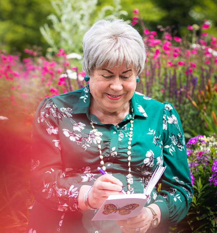 Pauline is looking down smiling as she writes in a small notebook. Behind her are pink and purple flowers. Pauline is a trans woman wearing a green dress with flowers on it, she has short silver hair