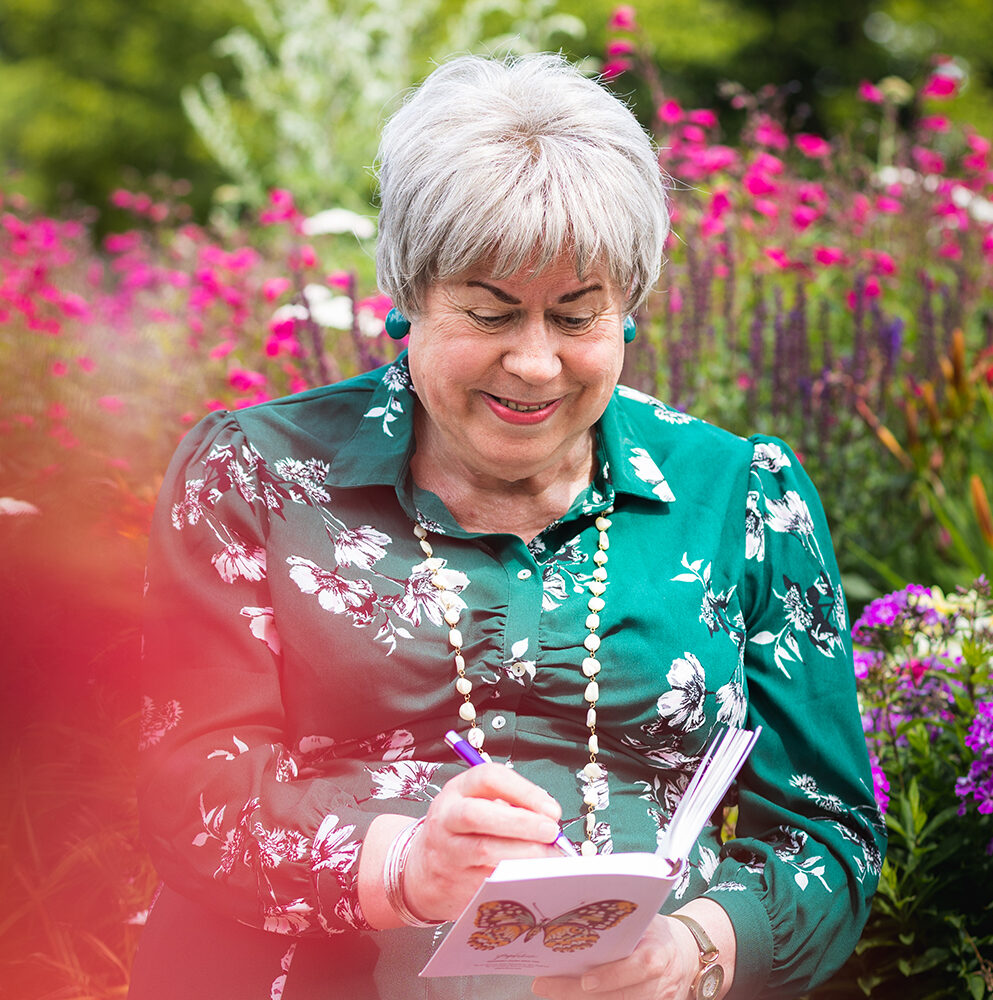 Pauline is looking down smiling as she writes in a small notebook. Behind her are pink and purple flowers. Pauline is a trans woman wearing a green dress with flowers on it, she has short silver hair