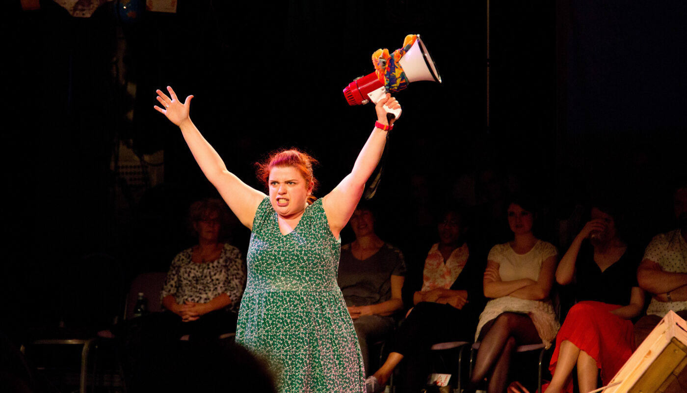 Sarah holds a loud hailer and outstretches both her arms above her head. Her teeth are gritted, as she performs in front of an audience in a darkened studio theatre.