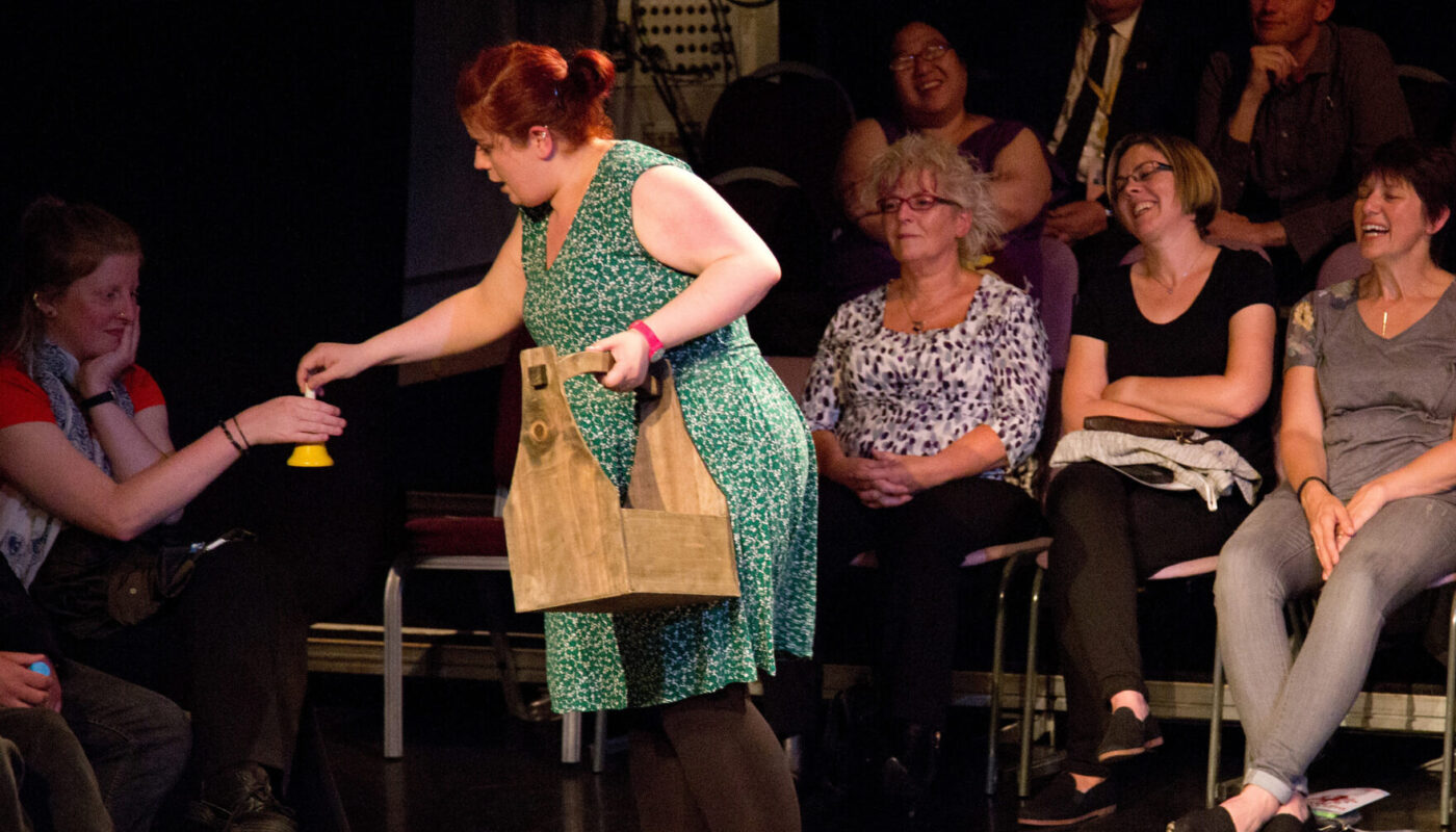 Sarah is barefoot onstage in a studio theatre. She hands a small yellow bell to an audience member while the rest of the audience look on.
