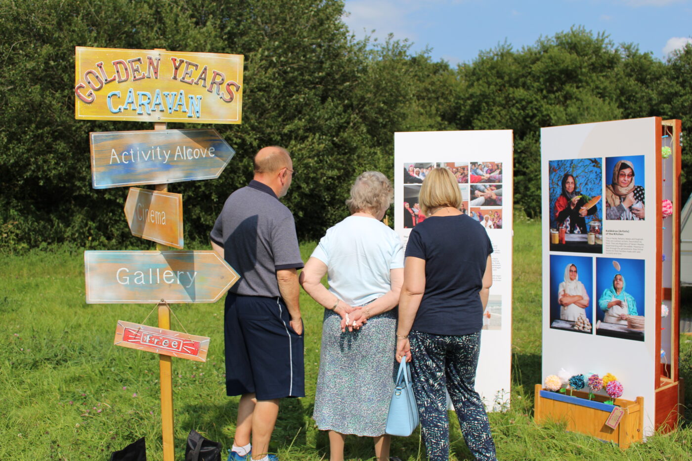 Three people stand in the sunshine looking at a display board of photography. Behind them, a sign post for Golden Years Caravan points the way to areas including Activity Alcove and Gallery.
