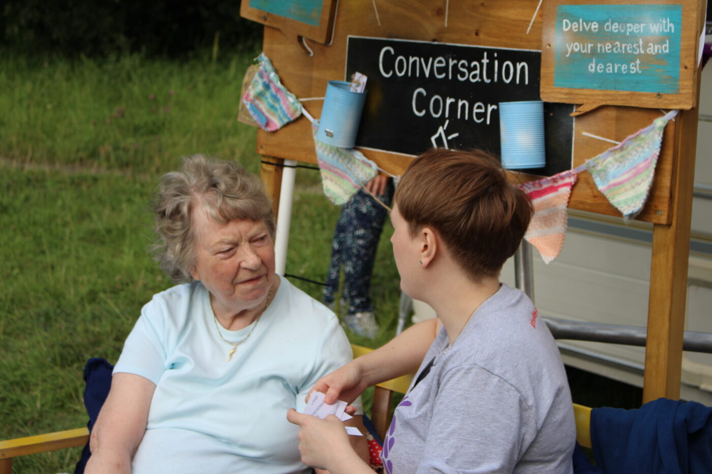Rachel sits on a bench chatting to an older person sitting next to them. A sign above their heads reads Conversation Corner.