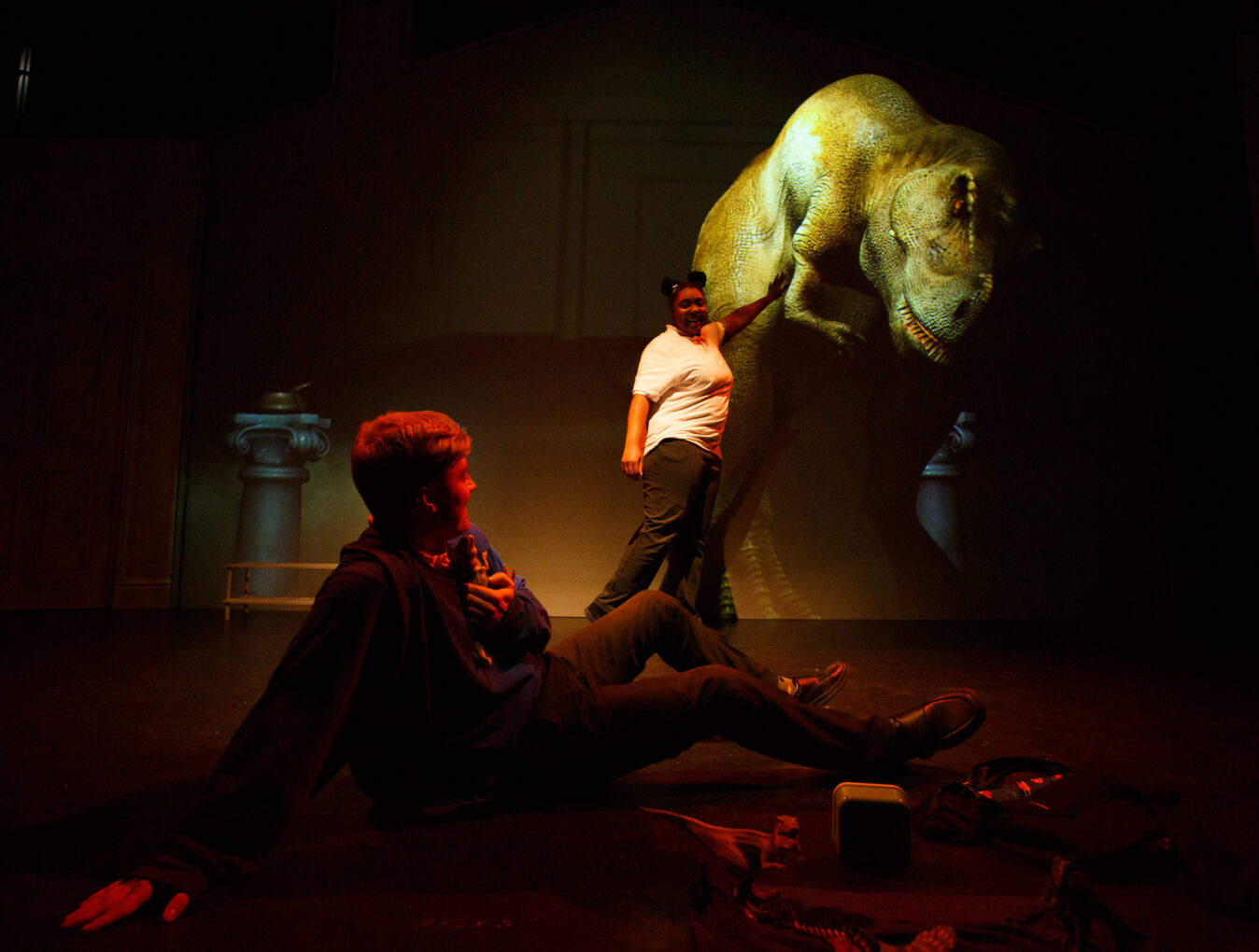 Two actors playing school children face a big projection of a T Rex. One of the actors touches the dinosaur, while the other cowers on the floor.