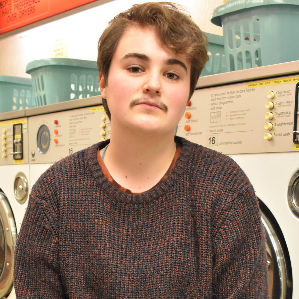 Roma is a young white person with short brown hair and a moustache. They sit in a launderette, wearing a knitted sweater.