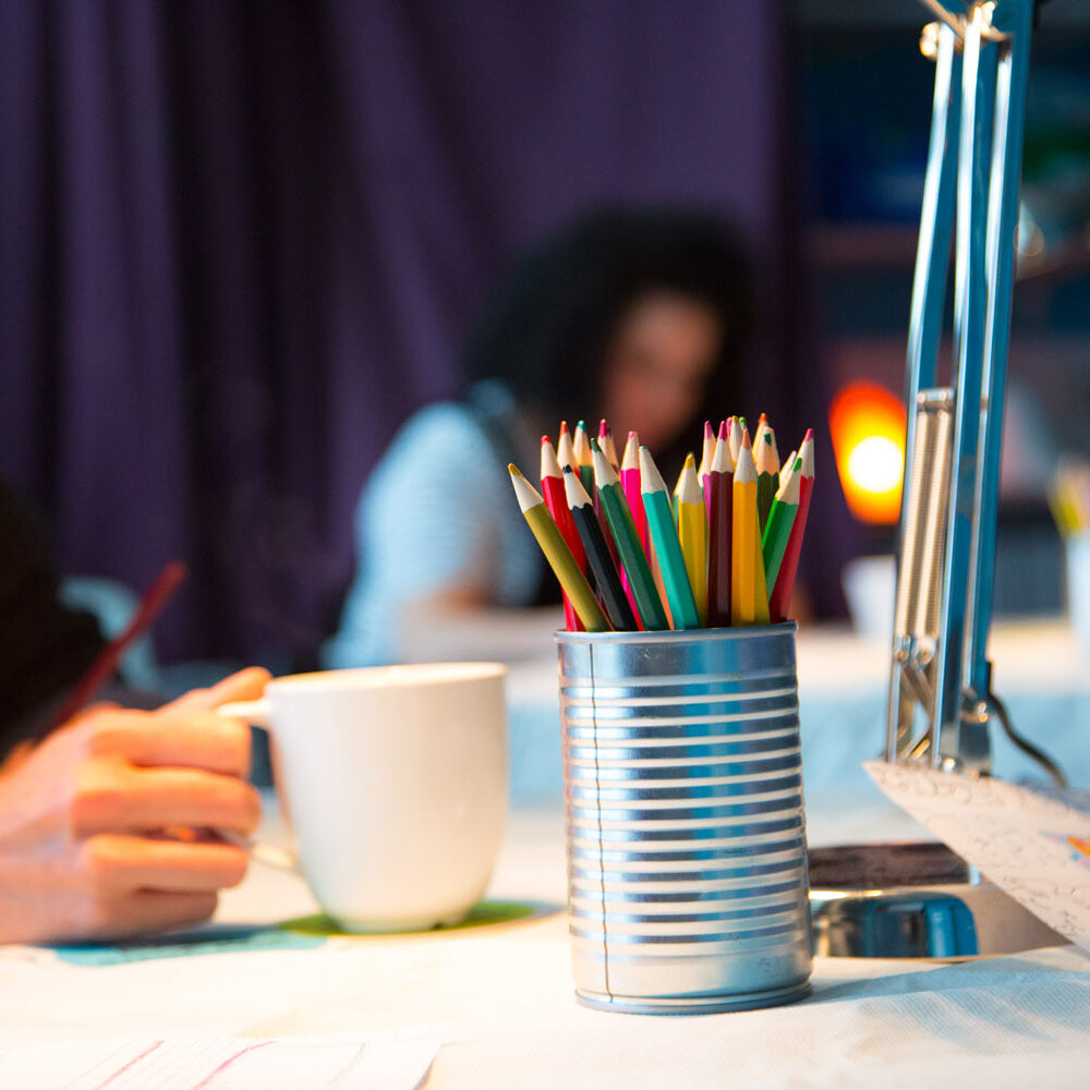 A pot of coloured pencils sits on a table. A person sits at the table with a mug in their hand.