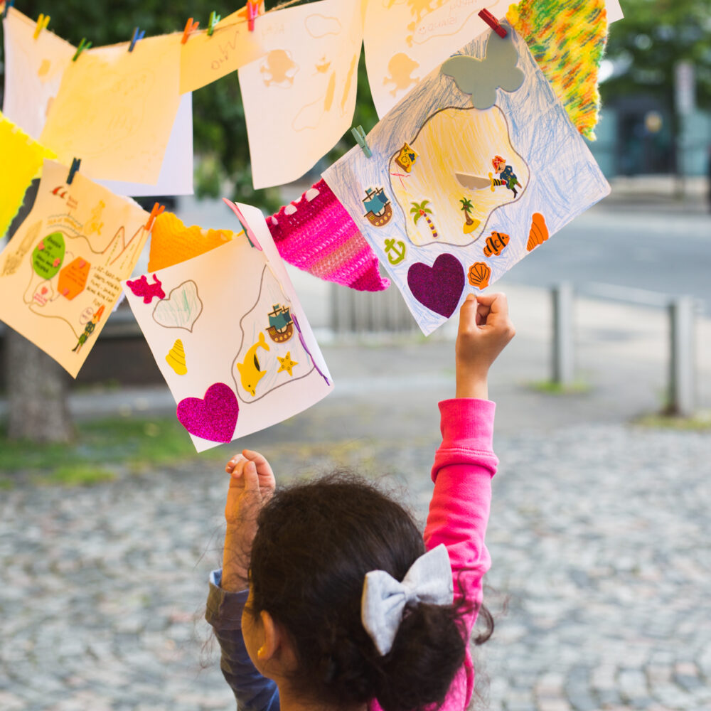 A young child reaches up to touch some drawings hung on a washing line.