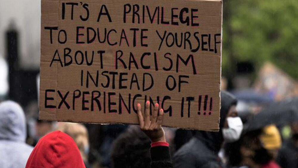 A protest sign reads 'It's a privilege to educate yourself about racism instead of experiencing it!!!'.