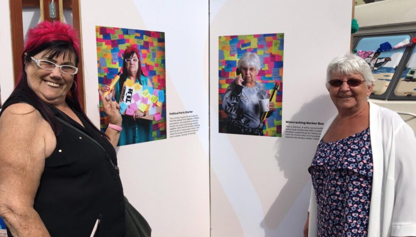 Two adults stand smiling beside photographic portraits of themselves.The portraits have bright multi-coloured backgrounds.