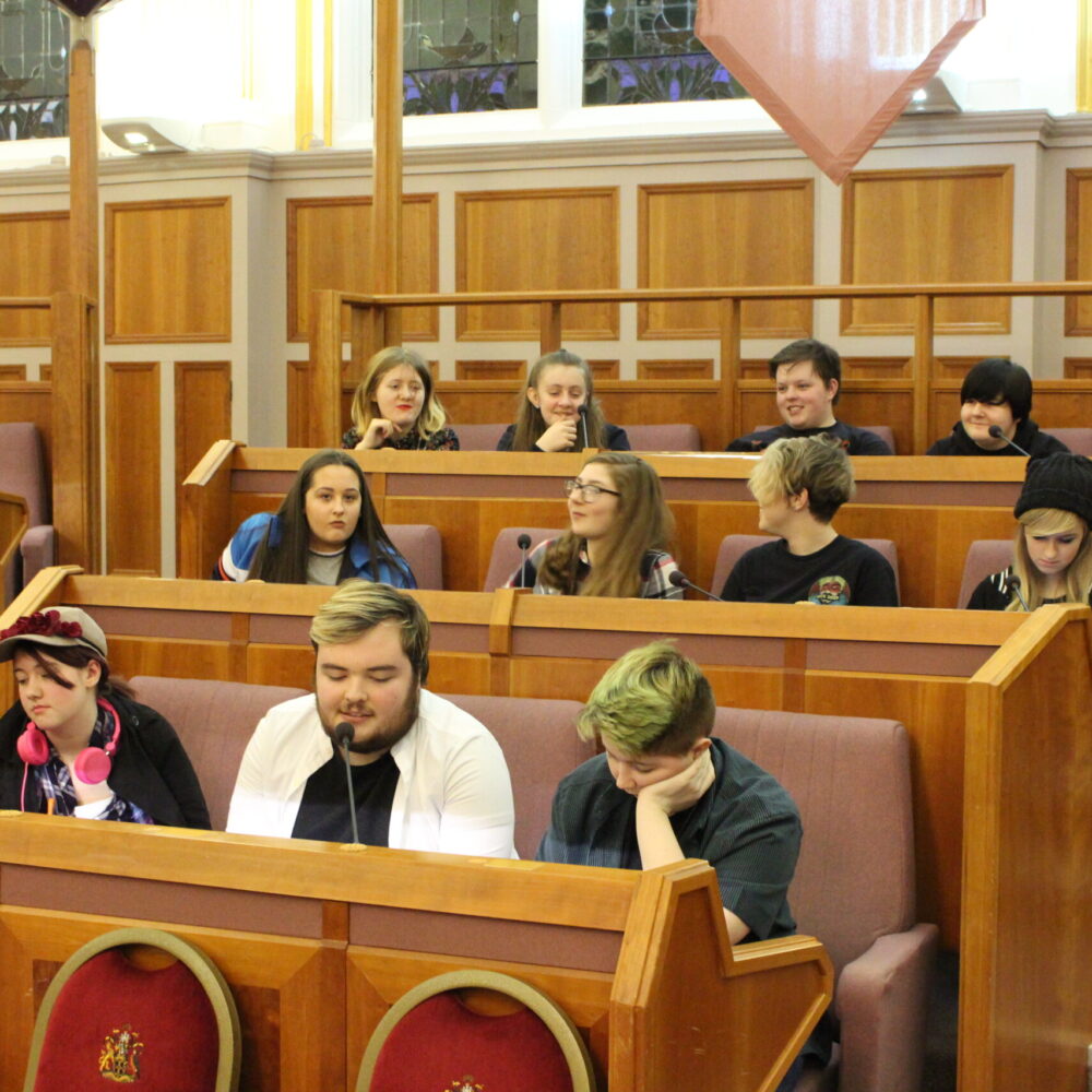 Twelve people sit in rows of a large civic chamber. The walls are panelled with wood and there is a microphone in front of each person. .