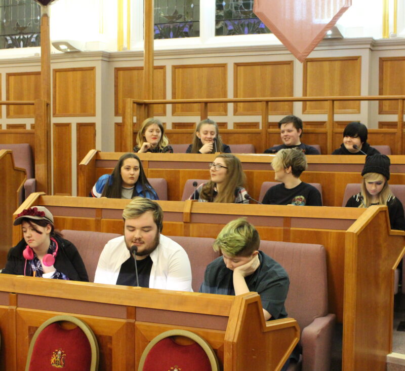 Twelve people sit in rows of a large civic chamber. The walls are panelled with wood and there is a microphone in front of each person. .