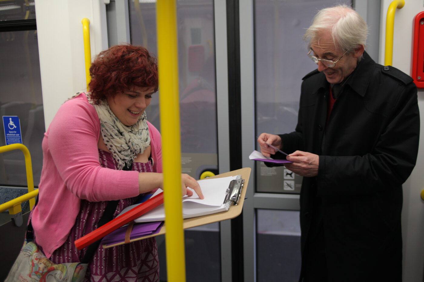 Sarah is on a tram looking at a clipboard and smiling. Next to her, a passenger holds a small card and smiles.