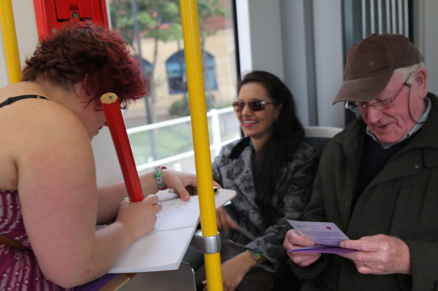 Sarah is on a tram using a giant pencil to a write in a notebook. Opposite her, two people are smiling.