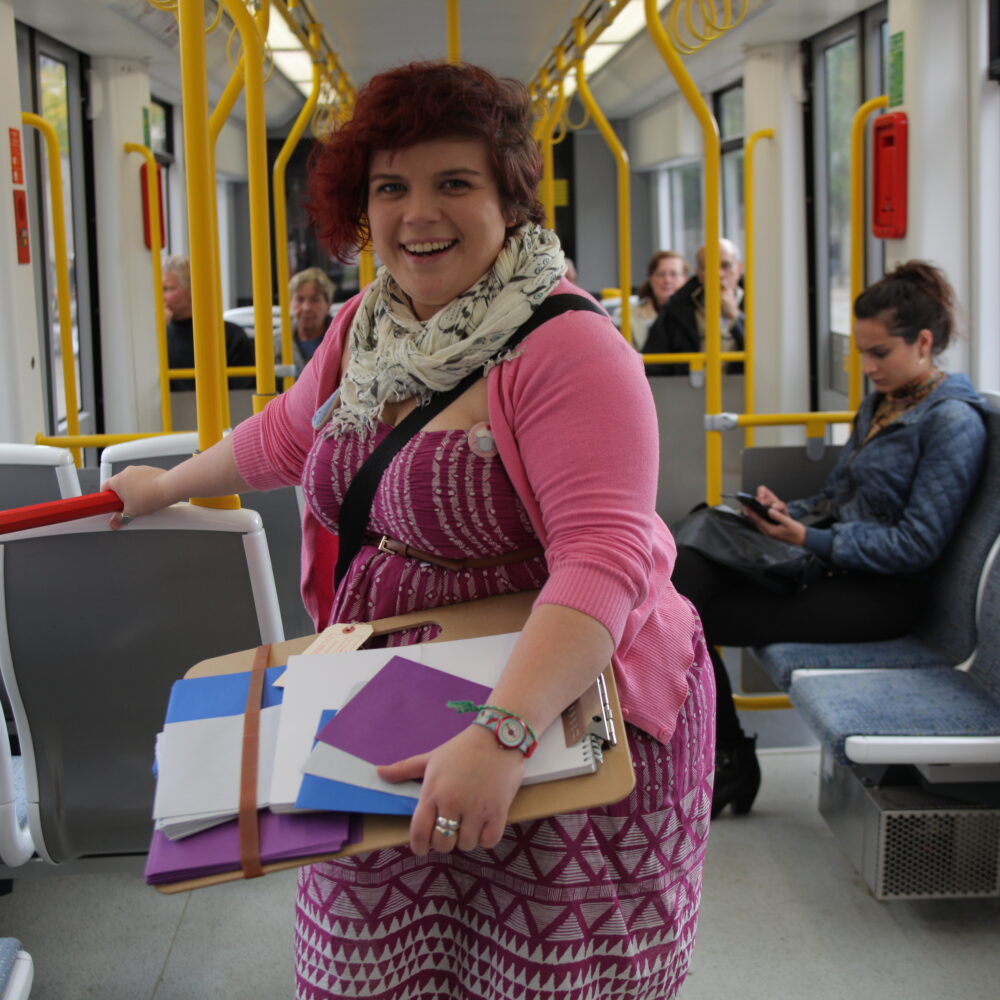Sarah stands in a tram carriage holding a giant red pencil and a clipboard. She is smiling.