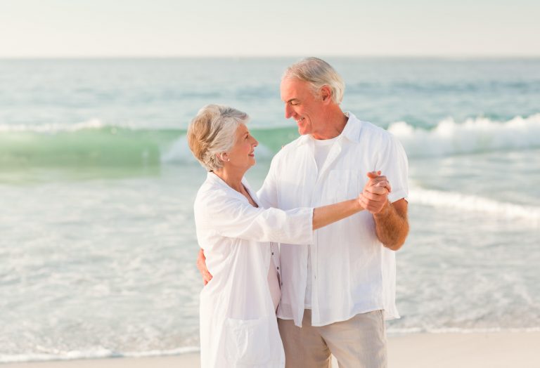 Photo of an older white man and woman dancing on a beach. They are dressed in white linen.