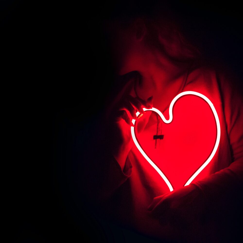 A person holds a red neon heart in their hands. The neon casts a red light on their body.