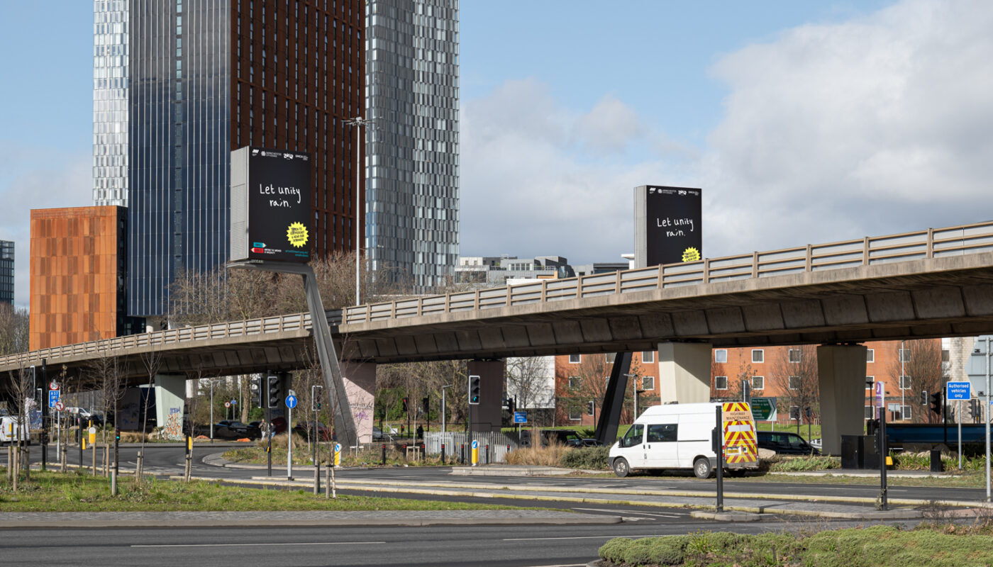Billboards on either side of the Mancunian Way feature children's writing that reads 'Let Unity Rain'.