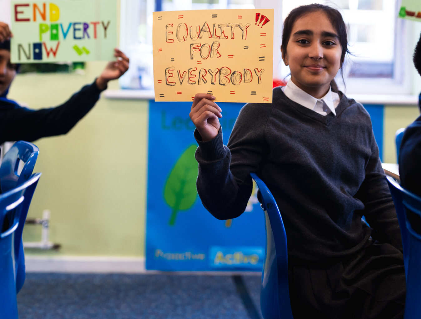 A year 6 pupil holds up a sign which says, 'Equality for Everyone'. In the background another pupil holds a sign reading 'End Poverty now'.