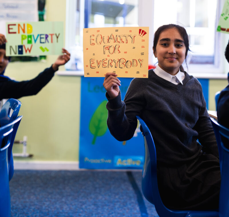 A year 6 pupil holds up a sign which says, 'Equality for Everyone'. In the background another pupil holds a sign reading 'End Poverty now'.