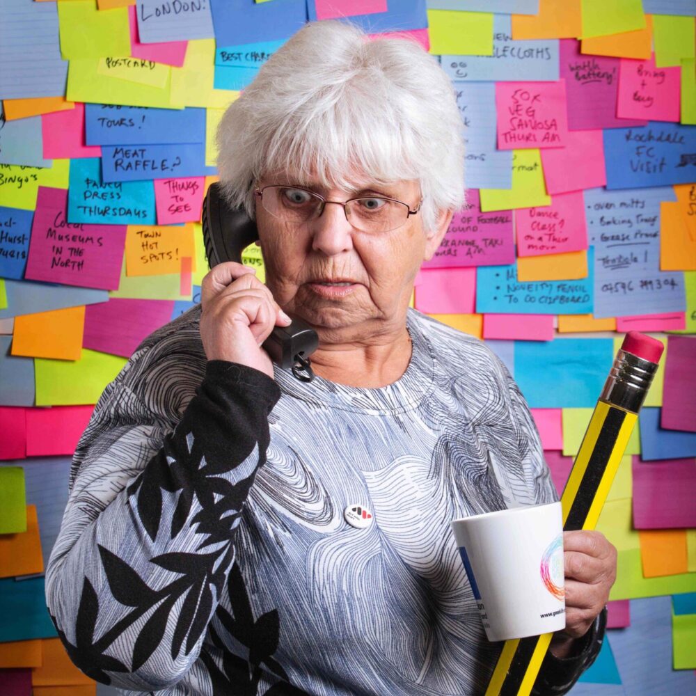 Denise, a white elder woman with white hair is stood on the telephone with a mug and pencil. Behind her is a wall of sticky notes.