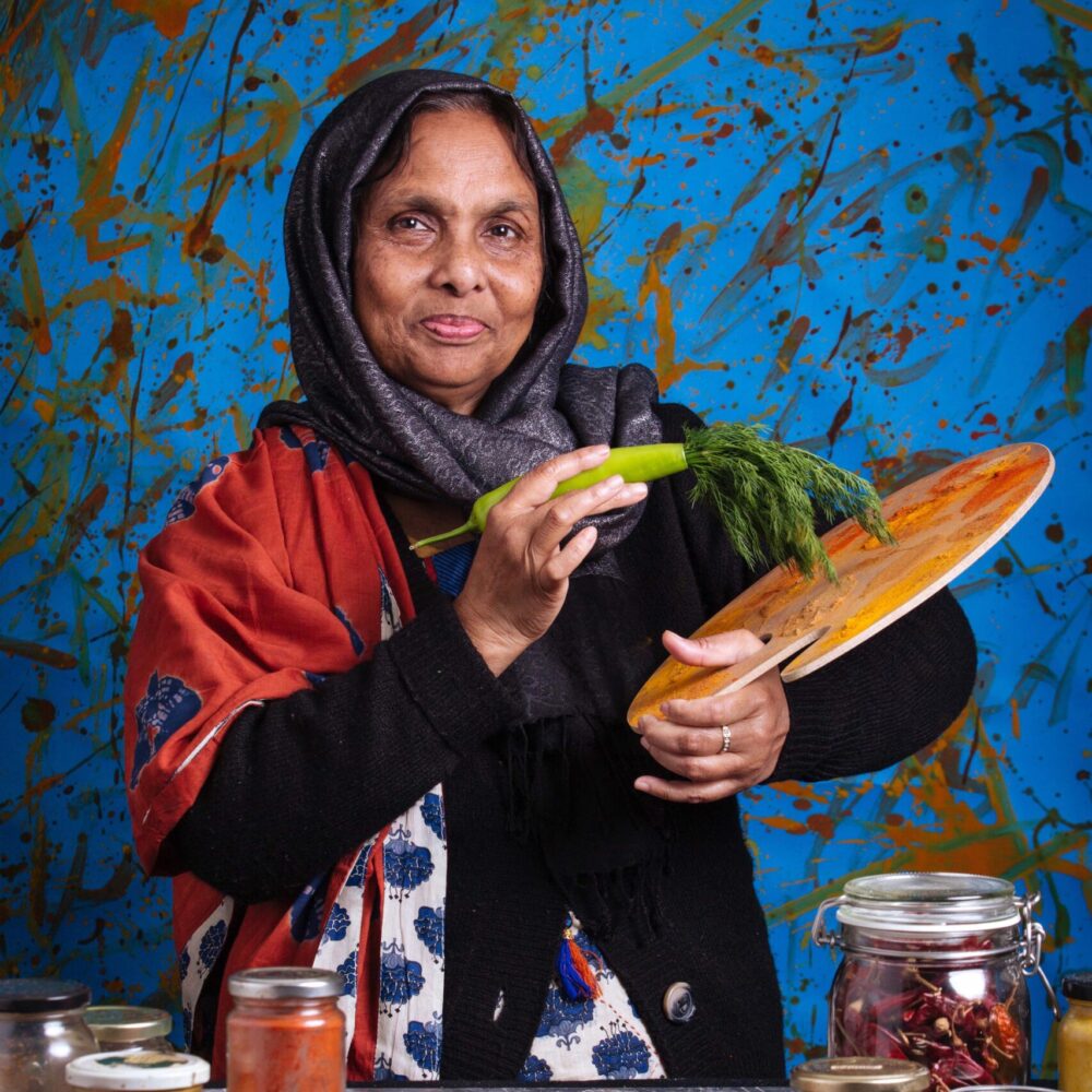 Farida, an elder South Asian woman, is stood looking dignified and holding a painting palette covered in spices. There are herbs and spices on the table in front of her and she's using a vegetable as a paintbrush. The background is covered in splashes of herbs and bright colours.
