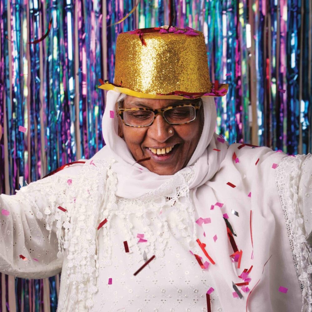 Nasim, an elder South Asian woman, is dressed in all white and stood dancing in front of a glittery curtain. She's wearing a gold glittery hat.