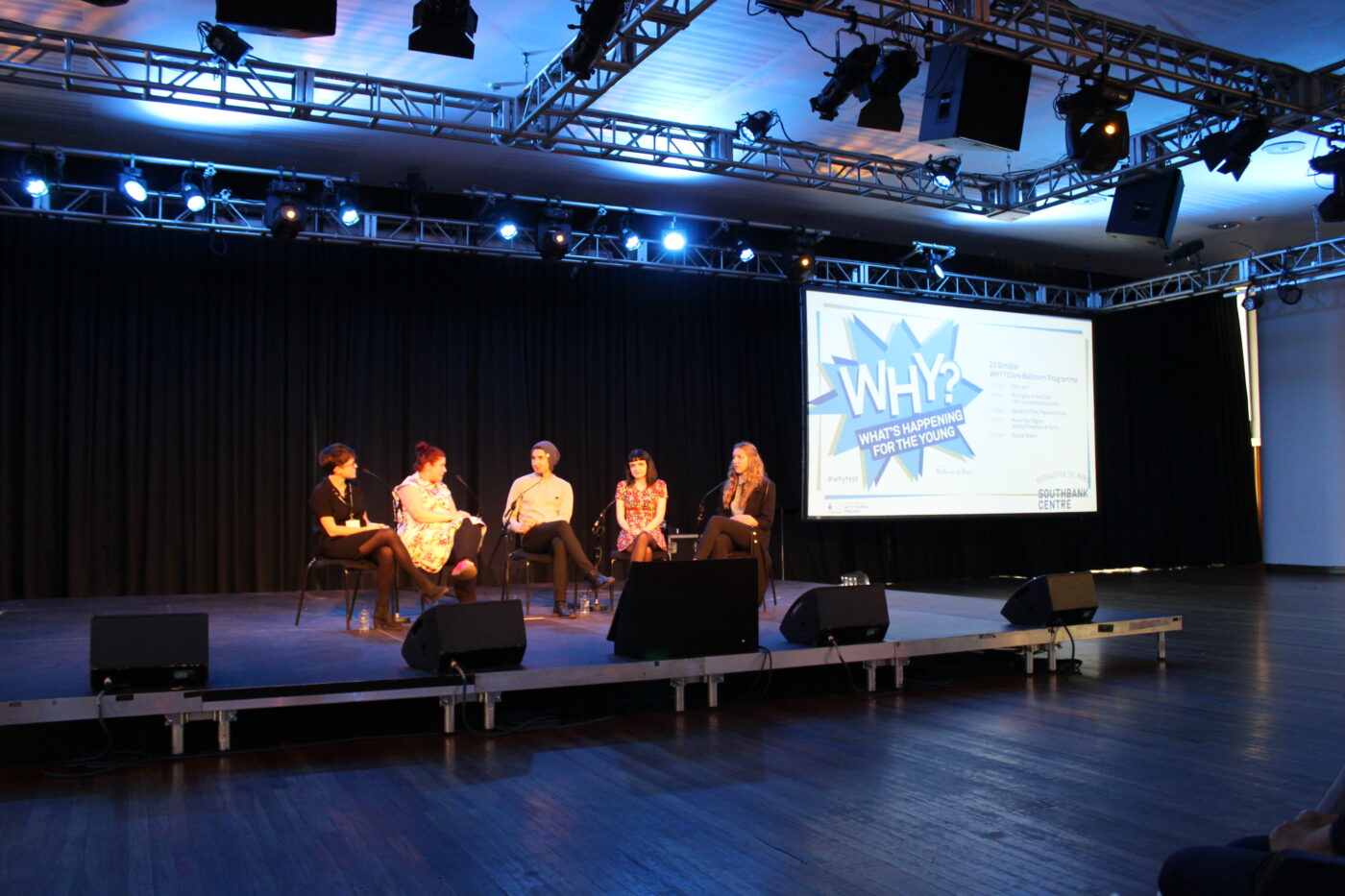 A panel of people on a stage speaking to each other, a screen reads 'WHY? what's happening for the young' in the background