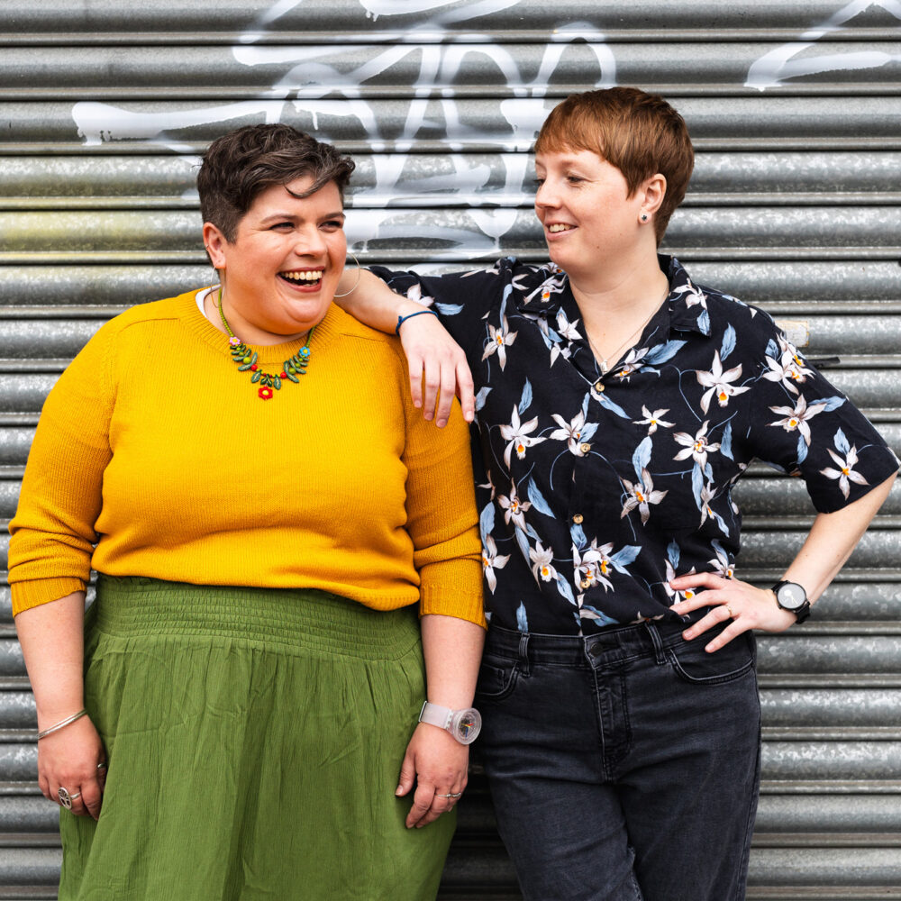 Sarah is looking into the distance and laughing as Rach leans on her shoulder and smiles.