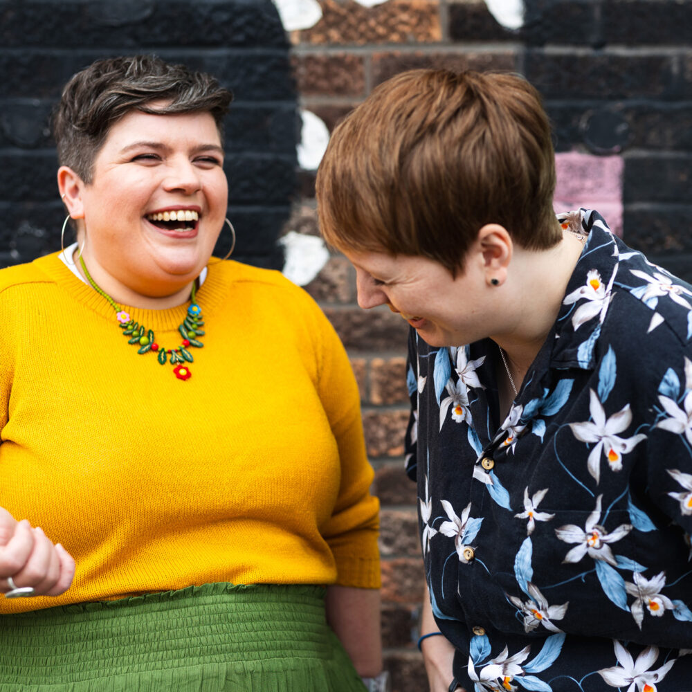 Sarah and Rachel are stood in front of a graffiti wall. Sarah is wearing a bright yellow jumper and floral necklace and laughing as she looks towards Rachel. Rachel's head is bowed slightly as she laughs, her eyes scrunched shut. She's wearing a floral shirt.