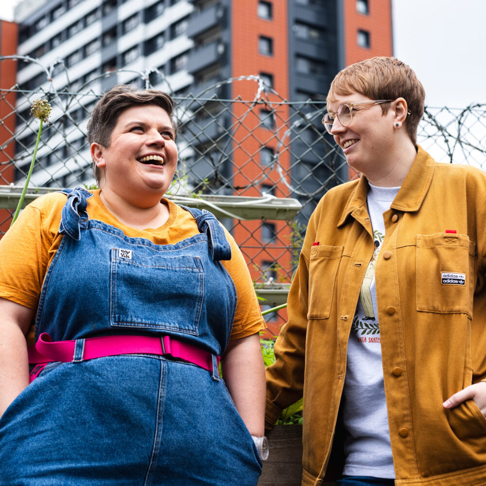 Sarah and Rachel are stood in front of a barbed wire fence. Sarah is wearing Lucy & Yak dungarees with her hands in he r pockets, grinning as she looks up. Rachel is wearing a yellow Adidas jacket and has a hand in her pocket, she's looking towards Sarah and smiling.