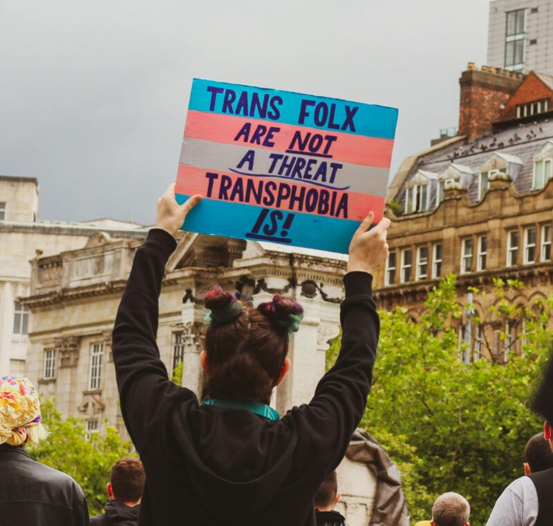 Someone at a protest holding a sign that says 'trans folx are not a threat, transphobia is!' 
