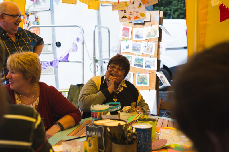 A woman sits at a craft table surrounded by people of all ages. She is laughing.