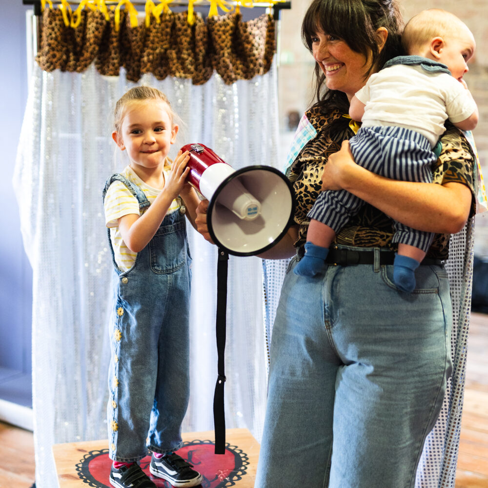 Image of a young child stood on a wooden box smiling, holding a megaphone whilst their parent holds their younger sibling.